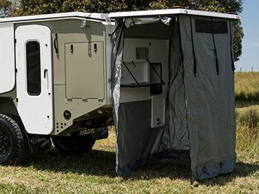 Off-road camper trailer with an open side door and a gray canvas privacy enclosure deployed over an outdoor kitchen area.