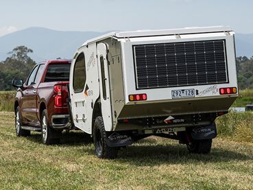 A red pickup truck tows a white camper trailer equipped with a large solar panel on its rear, parked on a grassy field.