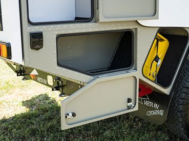 An open storage compartment on the side of a beige off-road camper trailer, showing a nearby yellow container.