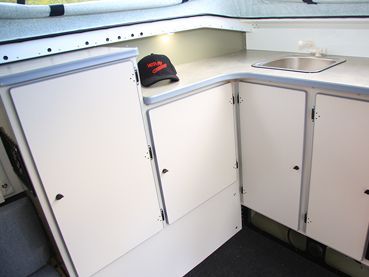The interior of a camper featuring a white kitchenette with a sink, counter space, cabinets, and a black cap on the counter.