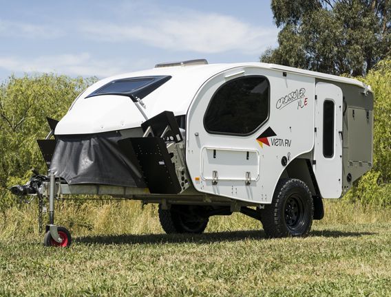 A white off-road camper trailer with black accents, parked on a grassy field under a blue sky.
