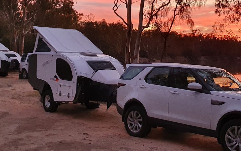 A white SUV tows a white teardrop-style camper with a pop-up roof at sunset near a lake.
