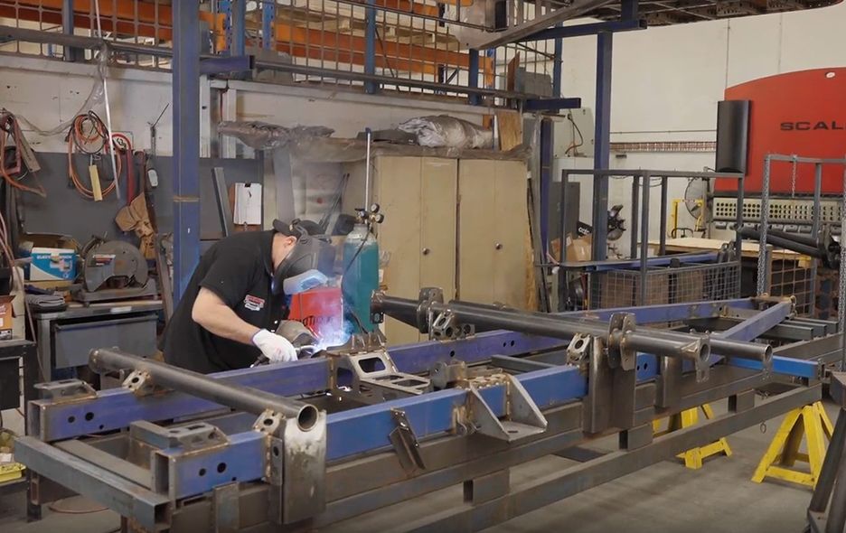 A person in a workshop wearing protective gear welds a large blue metal frame structure.