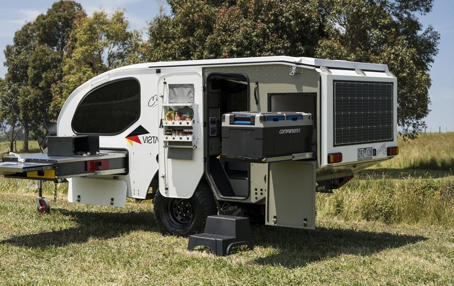 A white off-road camping trailer parked in a grassy field with its kitchen and storage compartments open.