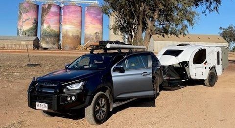 A black SUV towing a white camper trailer parked on a dirt lot in front of four large, mural-painted grain silos.