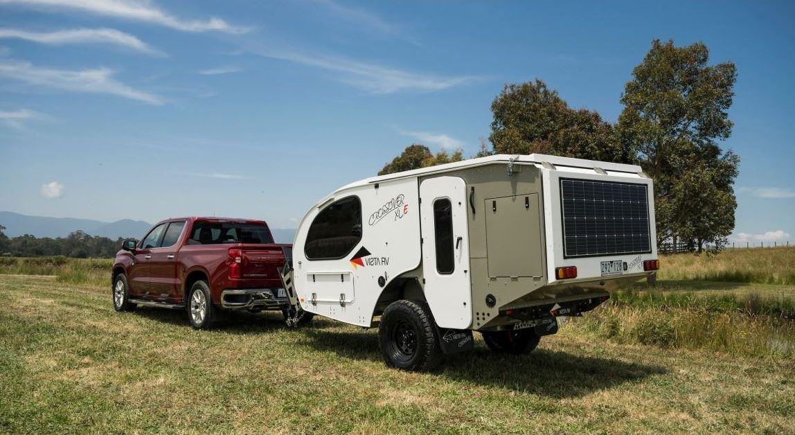 A red pickup truck tows a white teardrop-style camper across a grassy field under a bright blue sky.