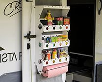 A white metal storage shelf mounted on a door, containing various pantry items and holding a roll of pink paper towels.