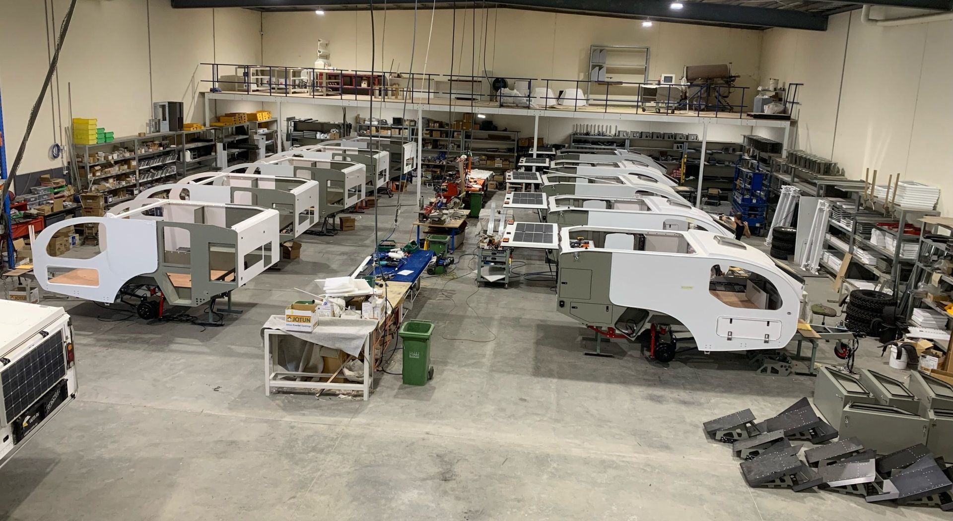 A wide-angle view of a workshop filled with multiple unfinished, white-paneled vehicle frames on a concrete floor.