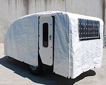 A teardrop camper covered by a white protective tarp with a window cutout and open door against a concrete wall.