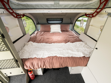 A cozy interior view of a camper van with a neatly made bed, featuring dusty rose bedding and windows on both sides.