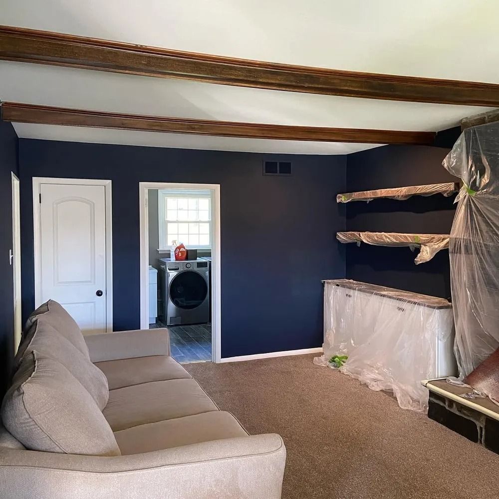 A living room with dark blue walls, wooden beams, and beige furniture, including a sofa. A white door and laundry room are visible.