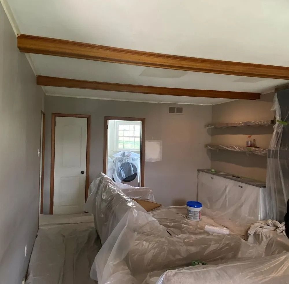 Room undergoing renovations with gray walls, stained wood beams, and furniture covered in plastic sheeting.