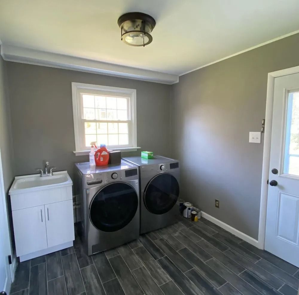 A laundry room with gray walls, dark wood-look floor tiles, and a washing machine and dryer. There's also a sink and an exterior door.