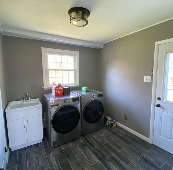 Laundry room with a washing machine, dryer, sink, window, and door. Gray walls, dark gray tiled floor.