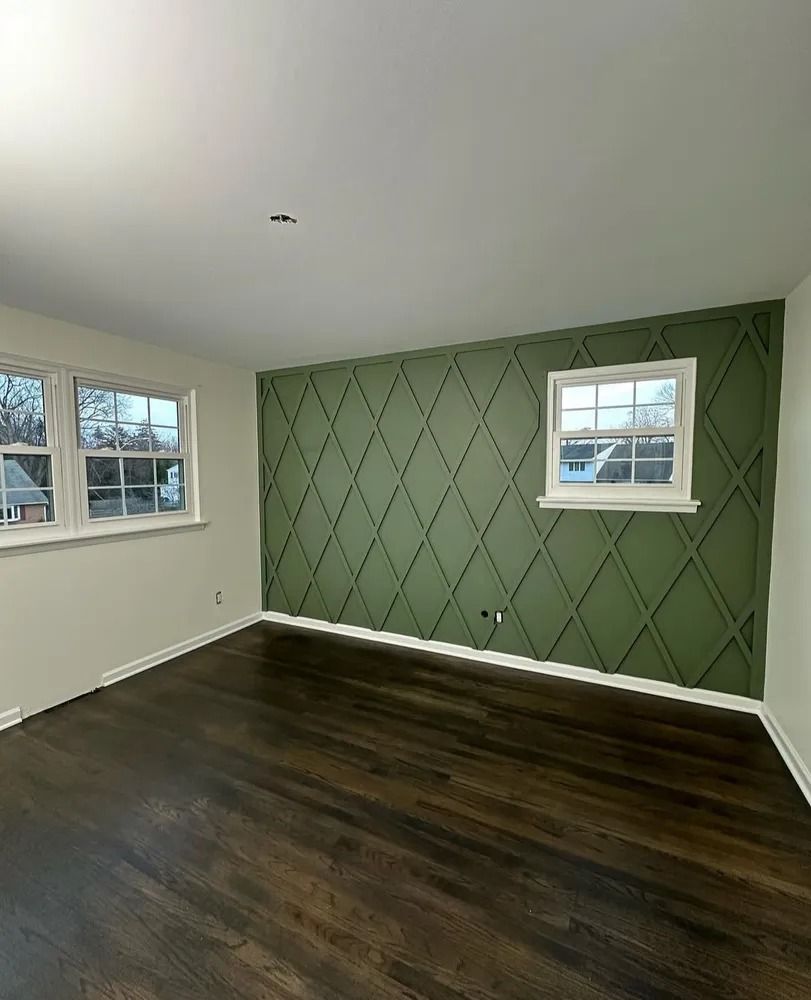 Empty room with dark wood floor, white trim and walls, one wall is olive green with a diamond pattern, two windows.
