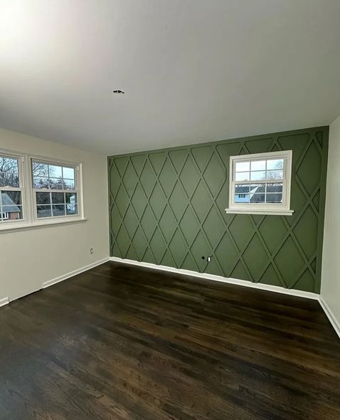 Empty room with dark wood floor, white walls, and a green diamond-patterned accent wall. Two white-framed windows.