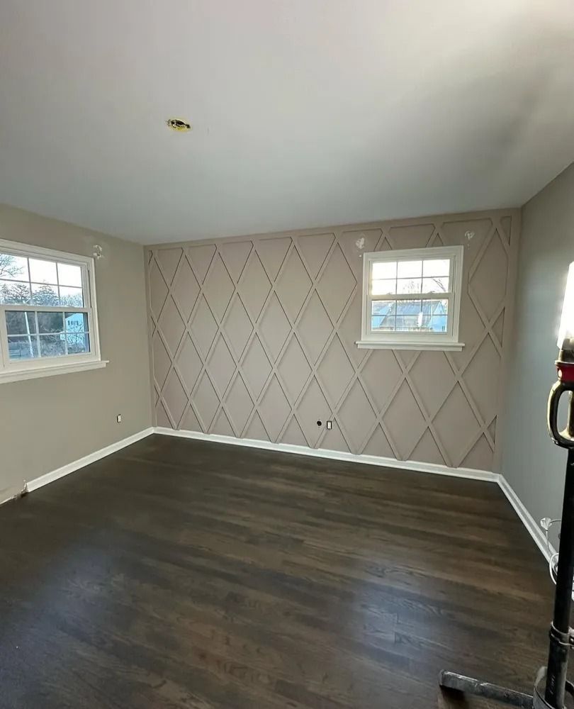 Empty room with dark wood floors, beige walls, and a diamond-patterned accent wall. Two windows provide natural light.