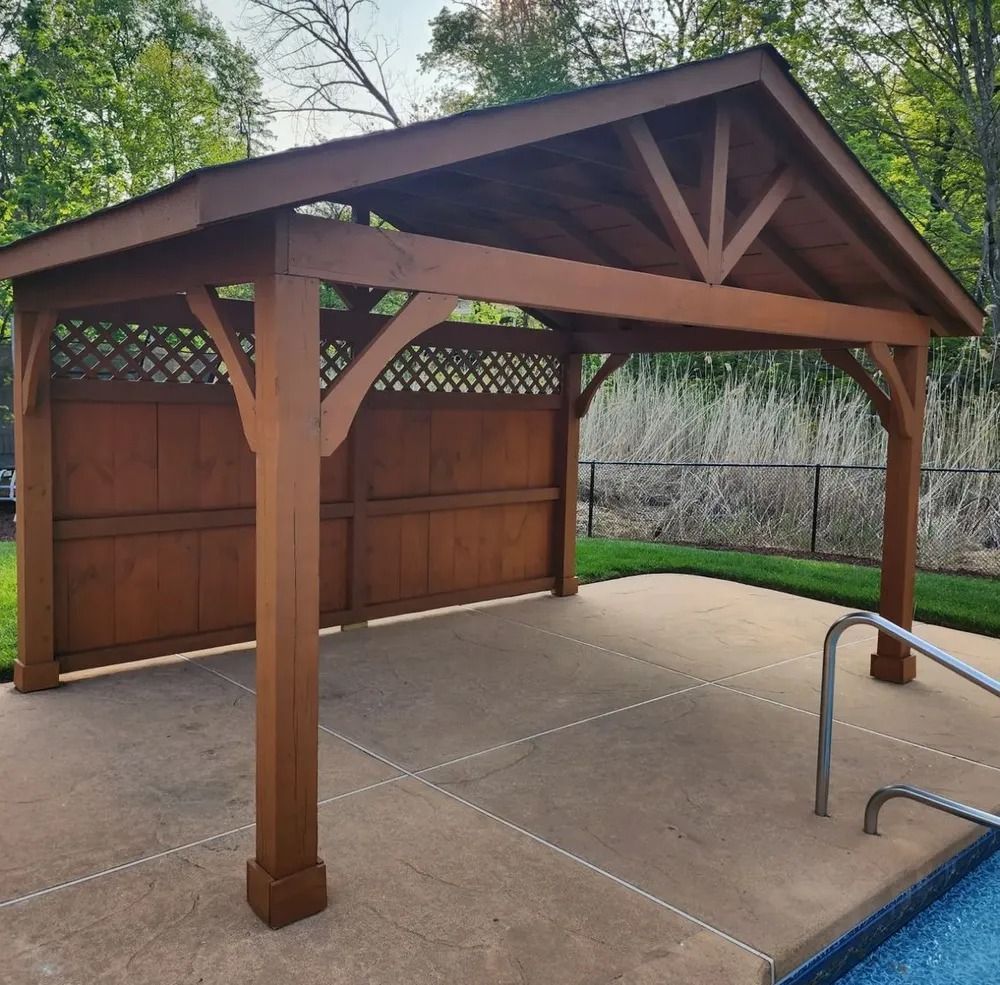 Wooden gazebo next to a pool. Brown structure with a sloped roof, latticework and a concrete patio.