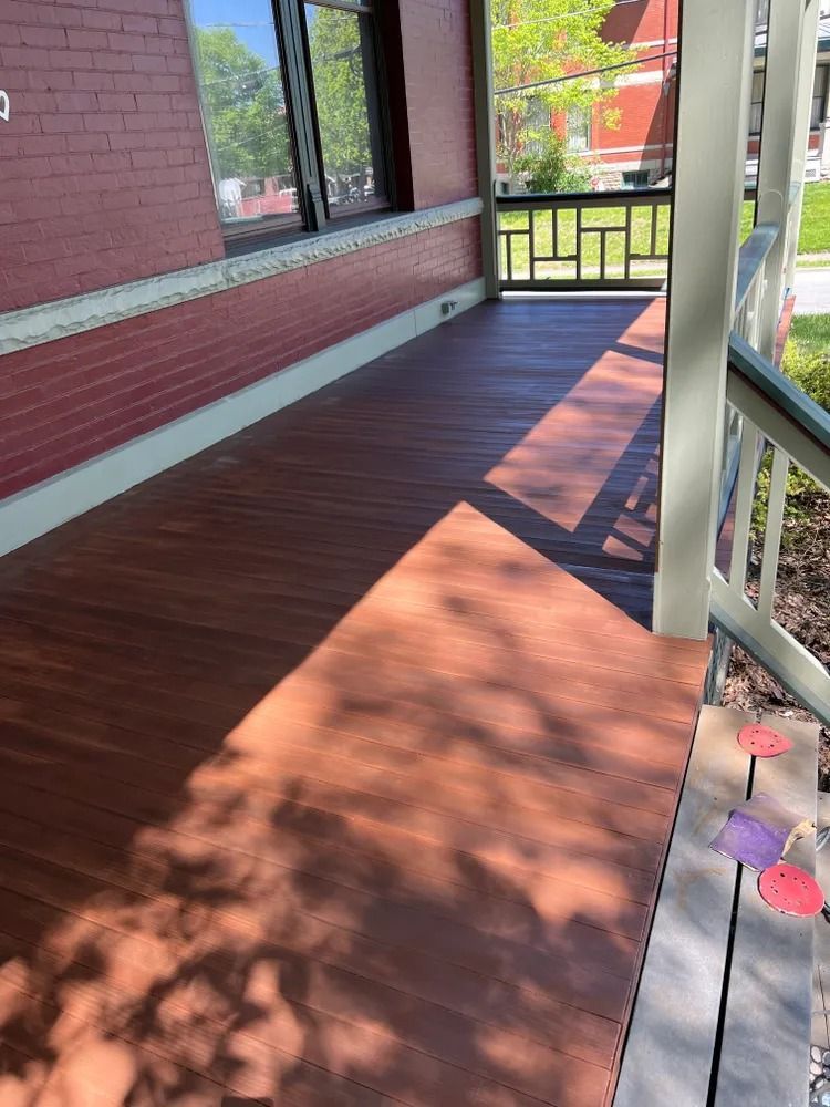 A wooden porch with a red brick wall, and green railing. Sunlight casts long shadows.