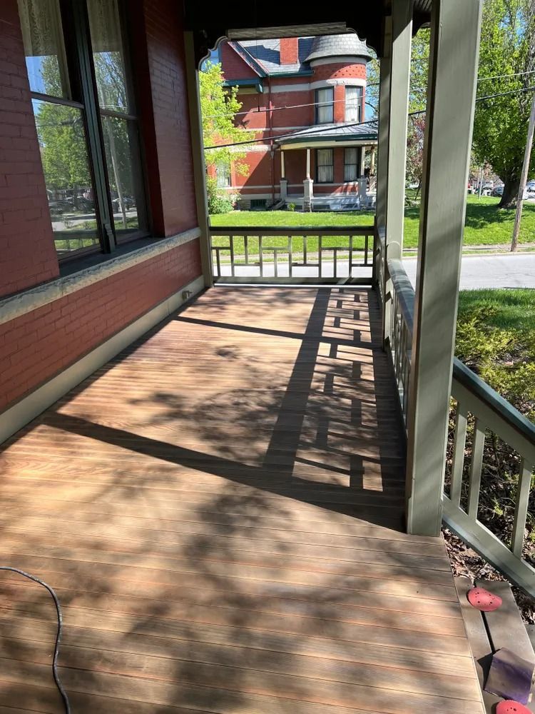 A wooden porch with brown decking and green support columns. A red brick building is on the left, with a view of houses and trees beyond the porch.