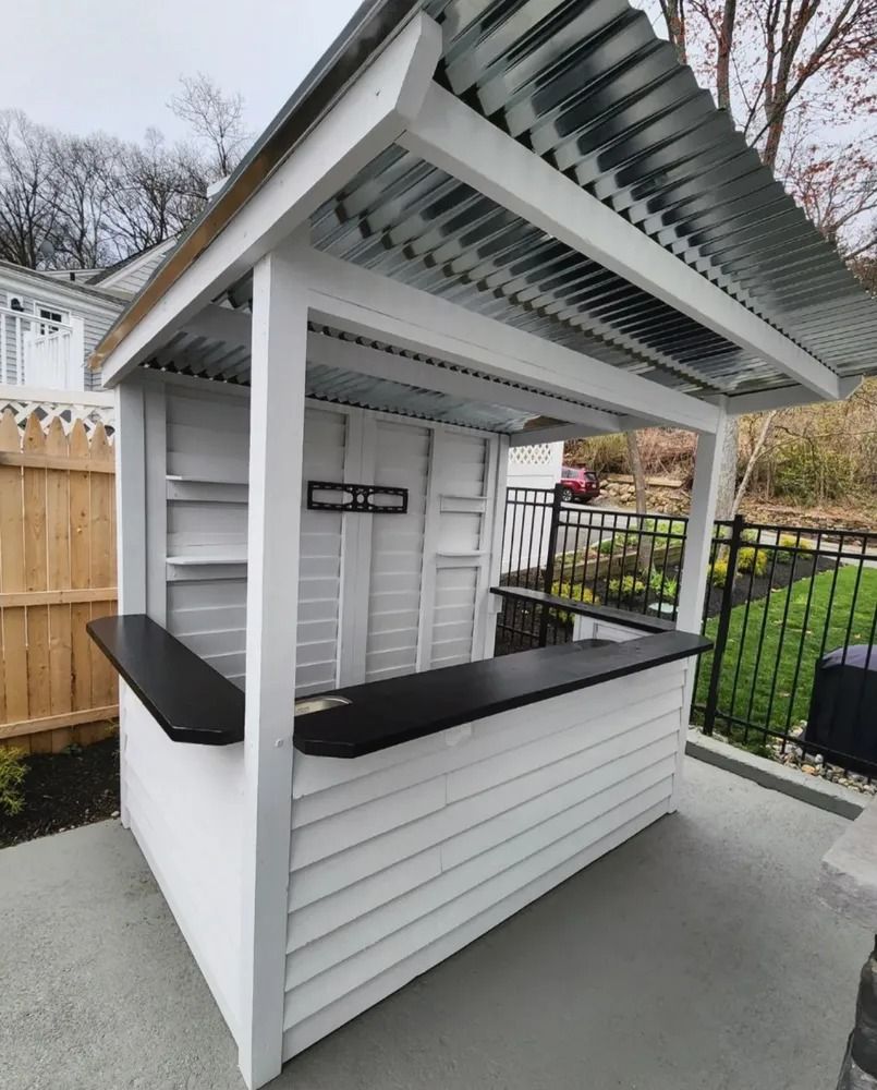 Outdoor white bar with black countertop and corrugated metal roof. Wooden structure is set on a patio near a fence and lawn.