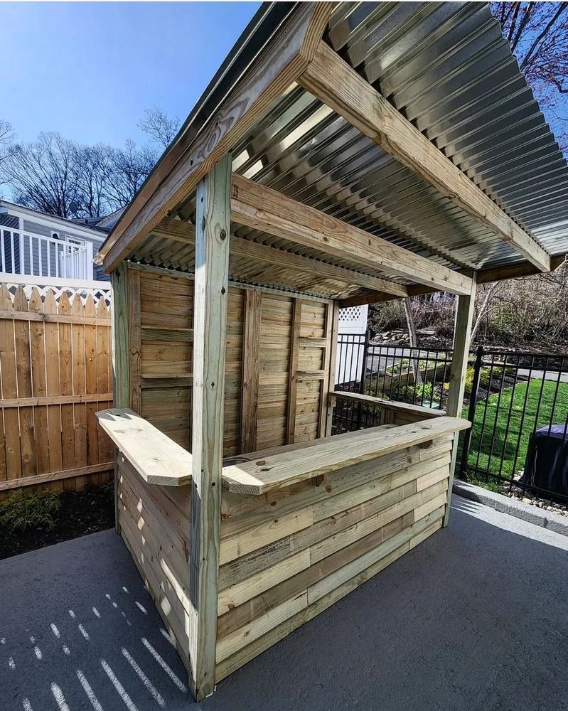 Outdoor wooden bar with corrugated metal roof on a patio. It has a counter and is constructed with raw wood.