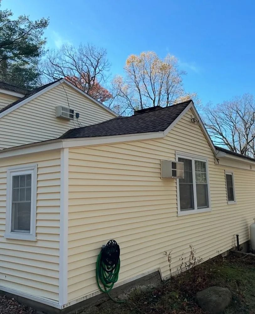 Yellow-sided house with a dark roof, white-framed windows, and a coiled green hose against the siding. Trees and a blue sky in the background.