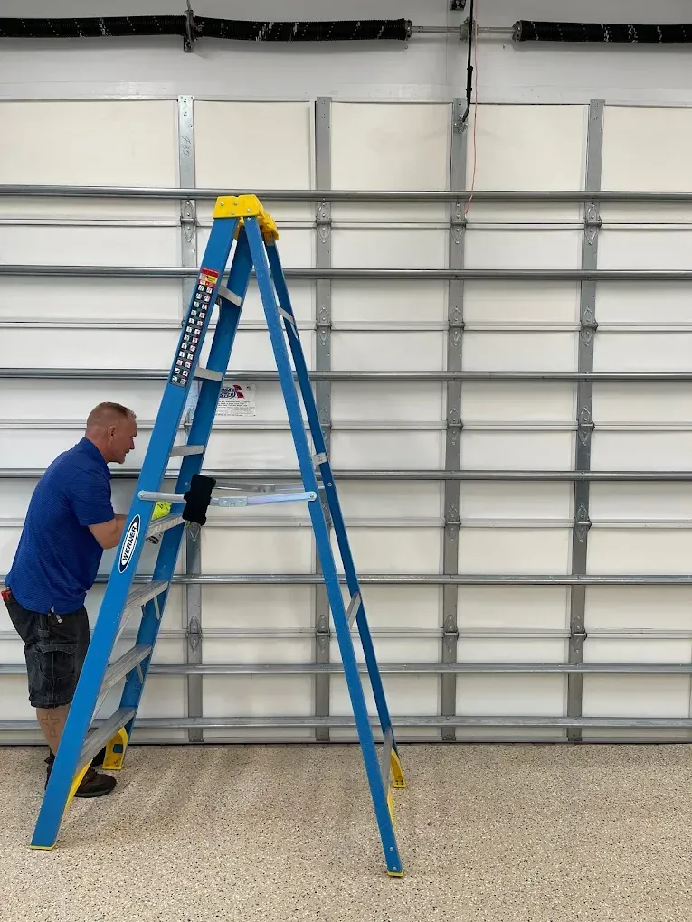 A person in a blue shirt stands on a blue stepladder, working on the interior of a white sectional garage door.