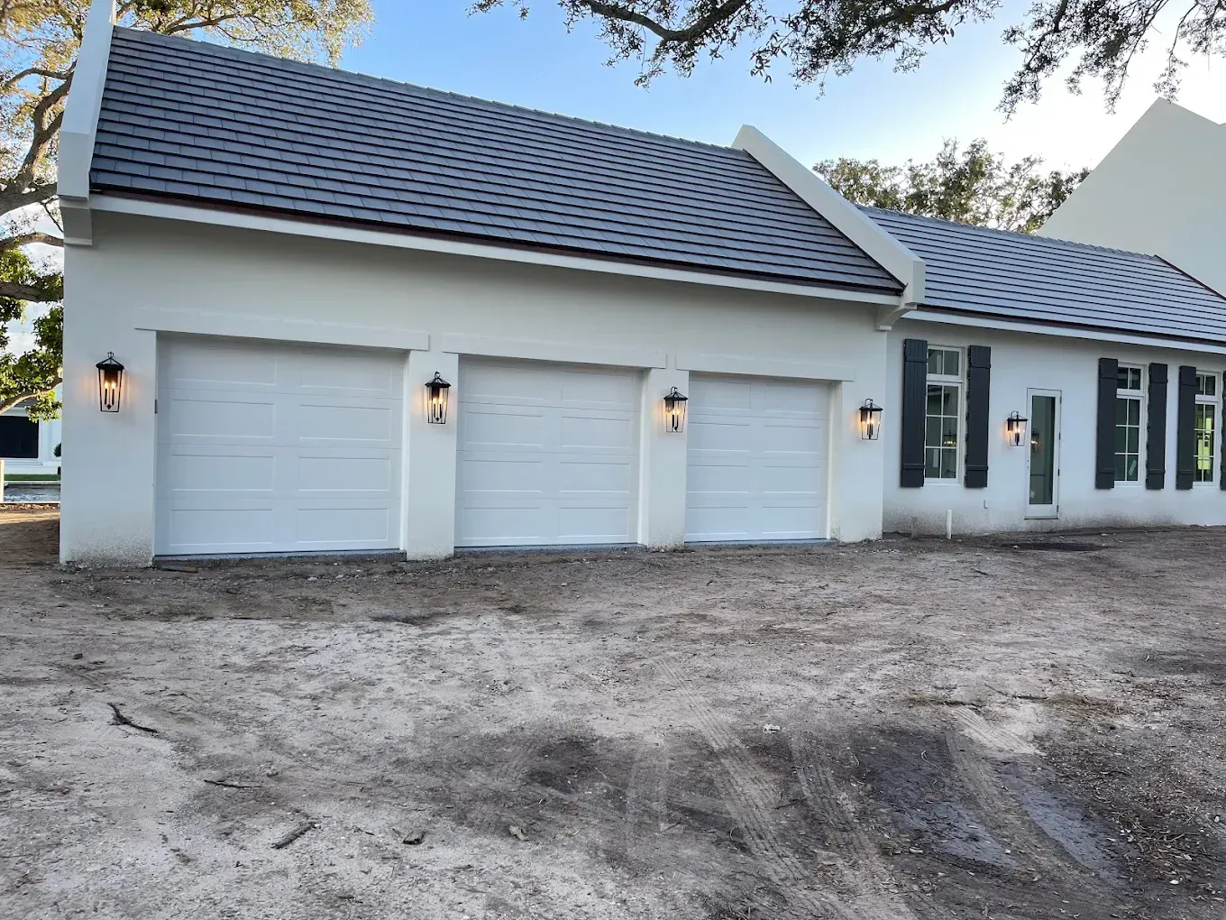 A white three-car garage building with a dark shingled roof, outdoor lanterns, and a dirt yard.