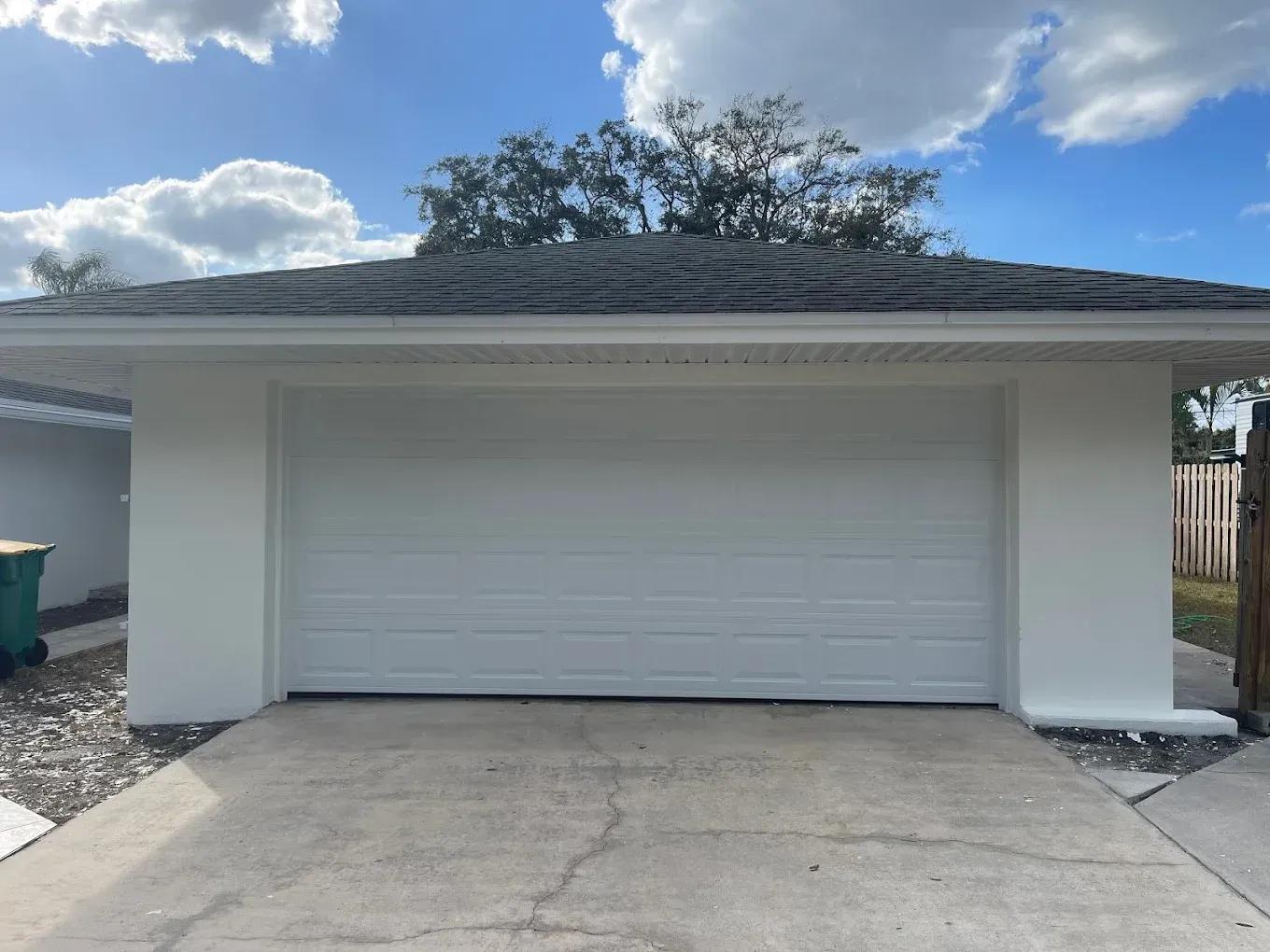 A white detached two-car garage with a dark gray shingled roof under a bright blue sky with scattered clouds.