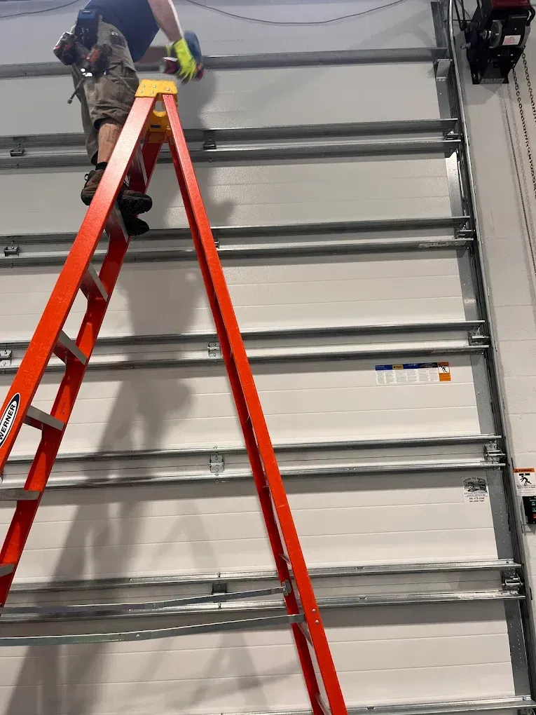 A person in work clothes stands on a tall orange ladder, installing hardware on a white paneled garage door.
