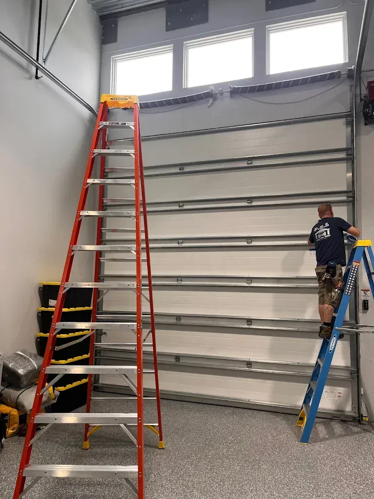 A worker stands on a blue ladder installing or repairing a white industrial garage door near a tall red ladder.