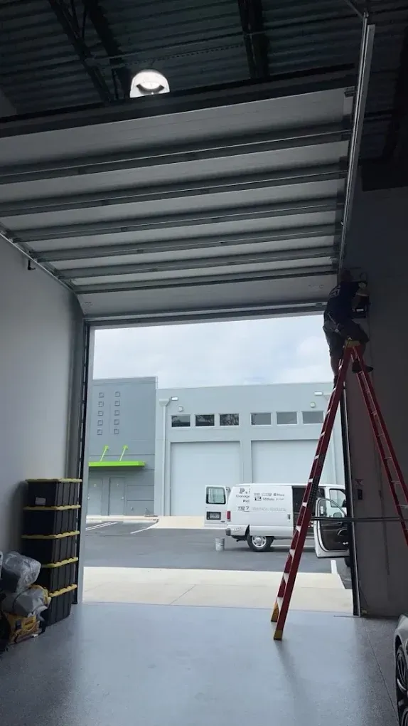 A person on a red ladder works on the track of an open industrial garage door, with a white van visible outside.