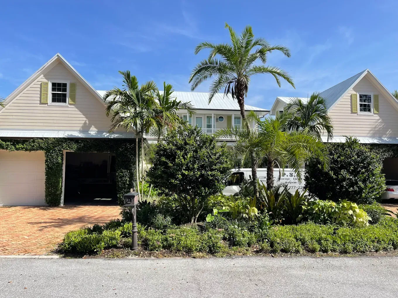 A light-colored two-story home with a metal roof, surrounded by tropical landscaping, palm trees, and a gravel driveway.