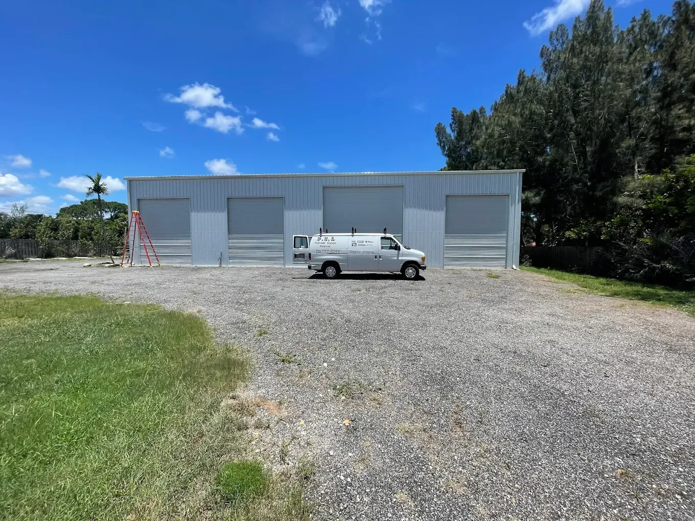 A white industrial warehouse with four garage doors and a work van parked in front on a gravel lot under a sunny sky.