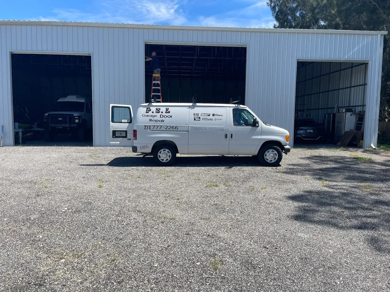 A white work van parked in front of a white metal industrial building with three open garage bays on a gravel lot.