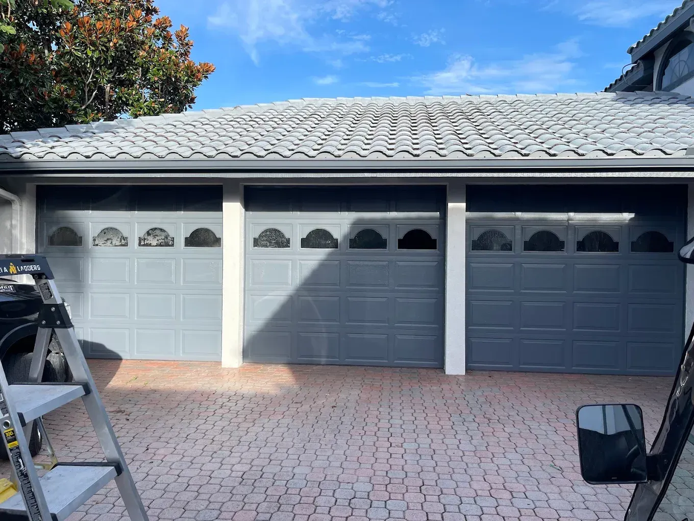 A three-car garage with gray paneled doors, arched window inserts, and a tiled roof, viewed from a paver driveway.