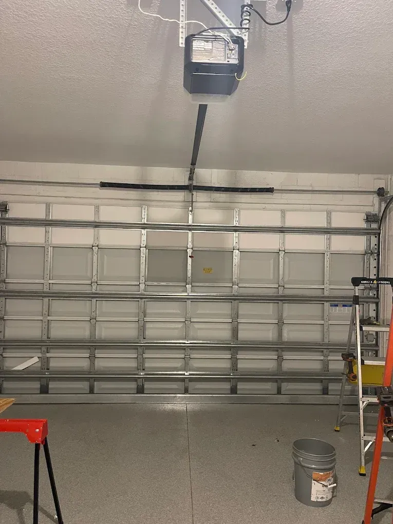 Garage door opener mounted on the ceiling above a closed white sectional garage door in a garage with speckled flooring.