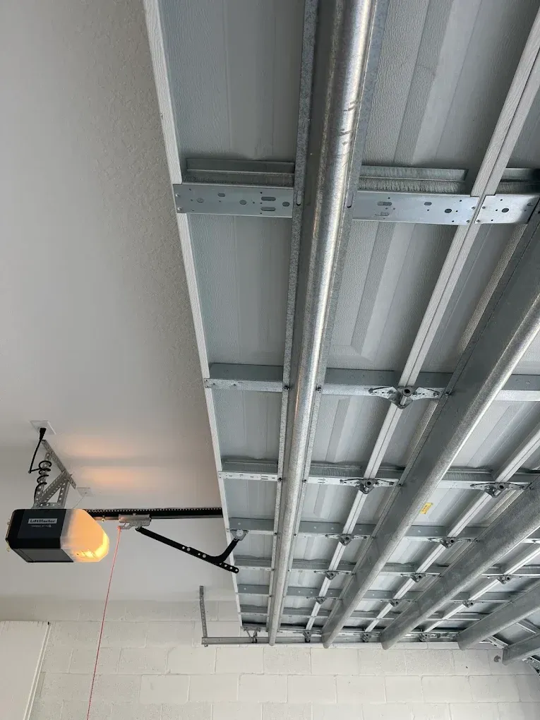 A garage door opener is mounted to a white ceiling, connected to the overhead metal tracks and panels of a garage door.