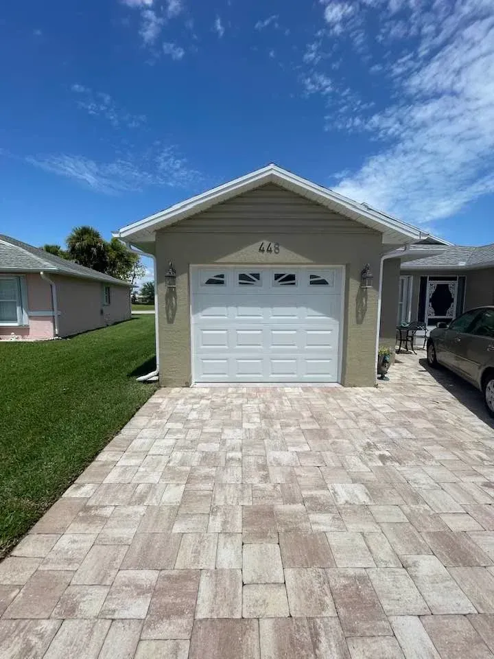 A tan, one-car garage with a white door and address 443 above, set in front of a paved driveway under a blue sky.