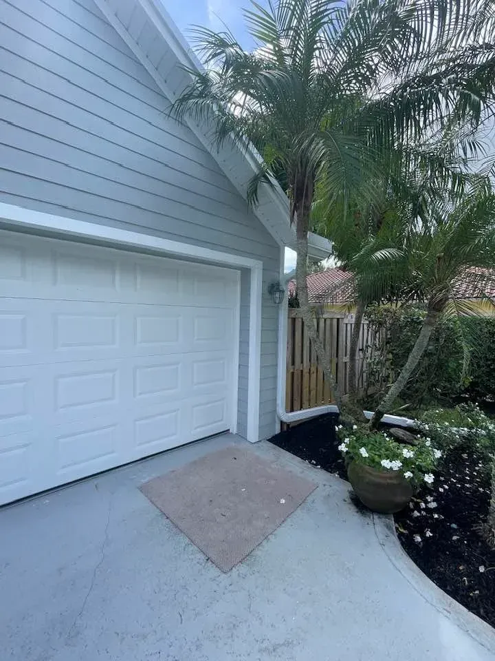 A gray-sided garage with a closed white door and a diamond-shaped concrete pad in a residential setting.