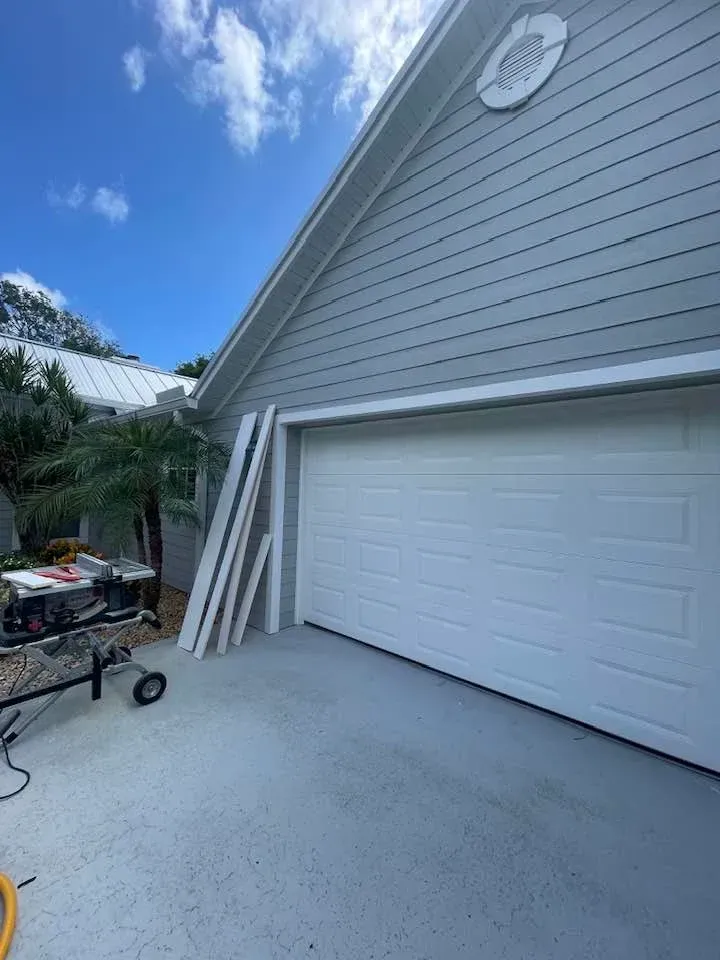 White garage door and gray siding on a house exterior, with construction materials and a table saw in the driveway.