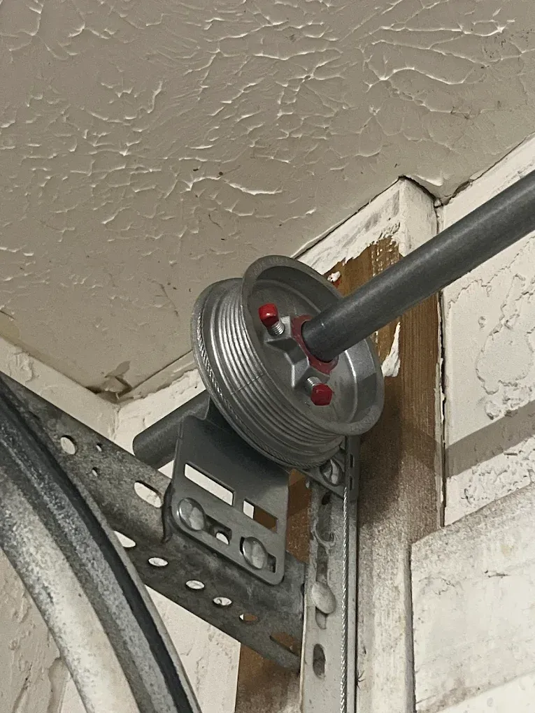 A metal cable drum attached to a garage door torsion bar mounted on a wall bracket.