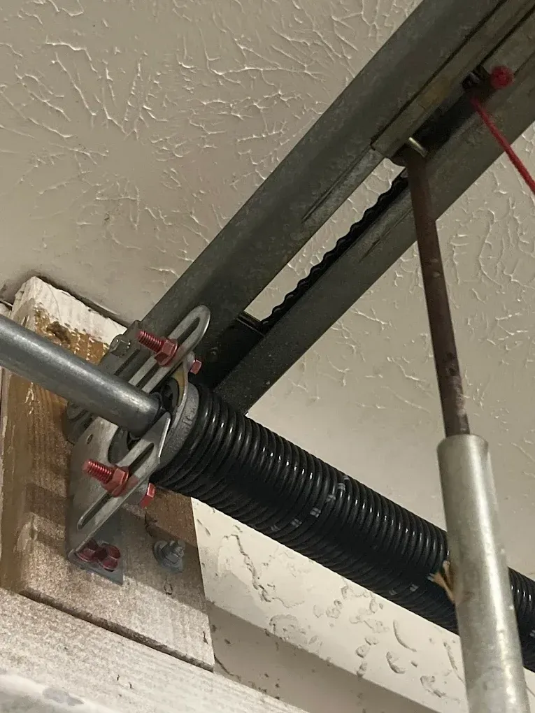 A close-up view of a black garage door torsion spring mounted on a metal shaft and wooden support beam near the ceiling.