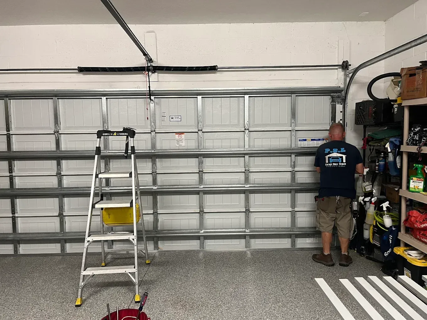 A technician in a garage works on a white metal sectional garage door with a metal ladder standing nearby.
