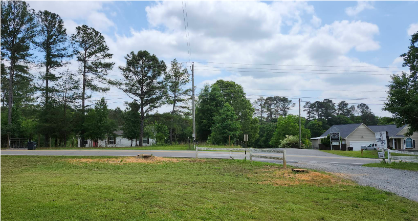 Green grassy field leads to a road with trees, houses, and power lines under a cloudy sky.
