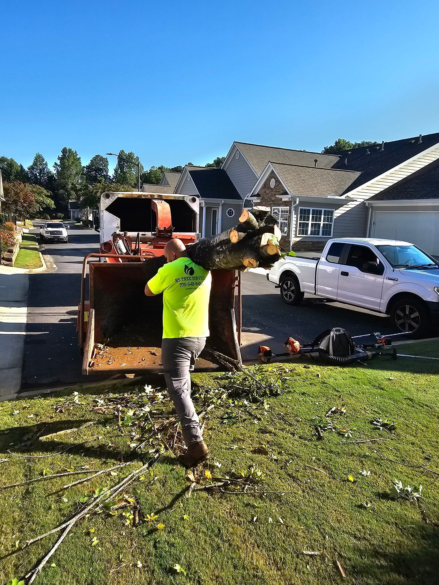 Man loading wood into a chipper in a residential yard. A white truck and houses are in the background.