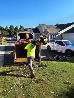 Man in neon green shirt, carrying logs to a wood chipper on a sunny day. Houses and a white truck in the background.