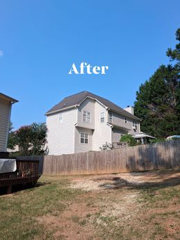 Two-story house with a gray roof and light-colored siding, behind a wooden fence, under a blue sky.