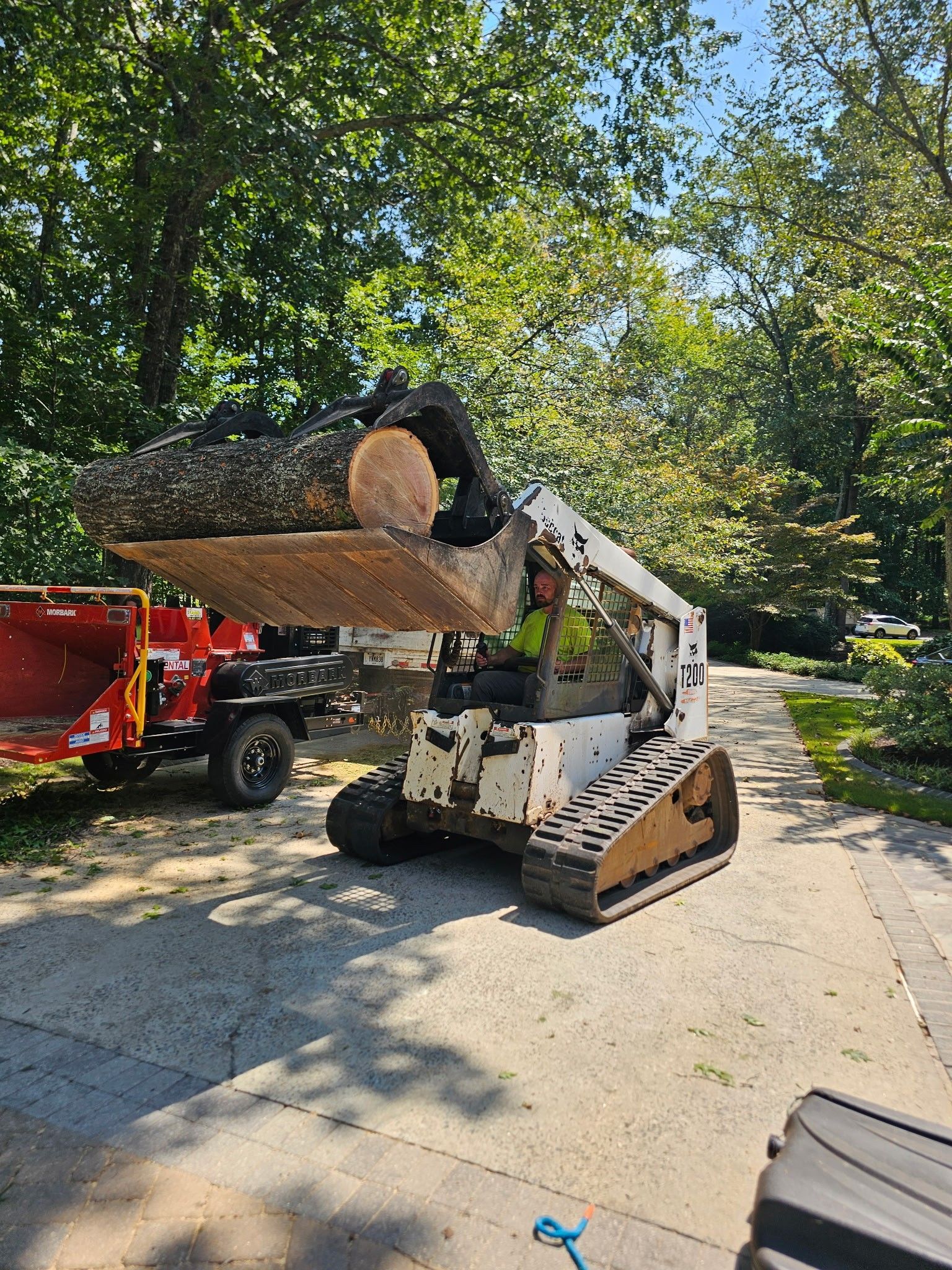Bobcat skid steer with log in its bucket, next to a wood chipper on a driveway, surrounded by trees.